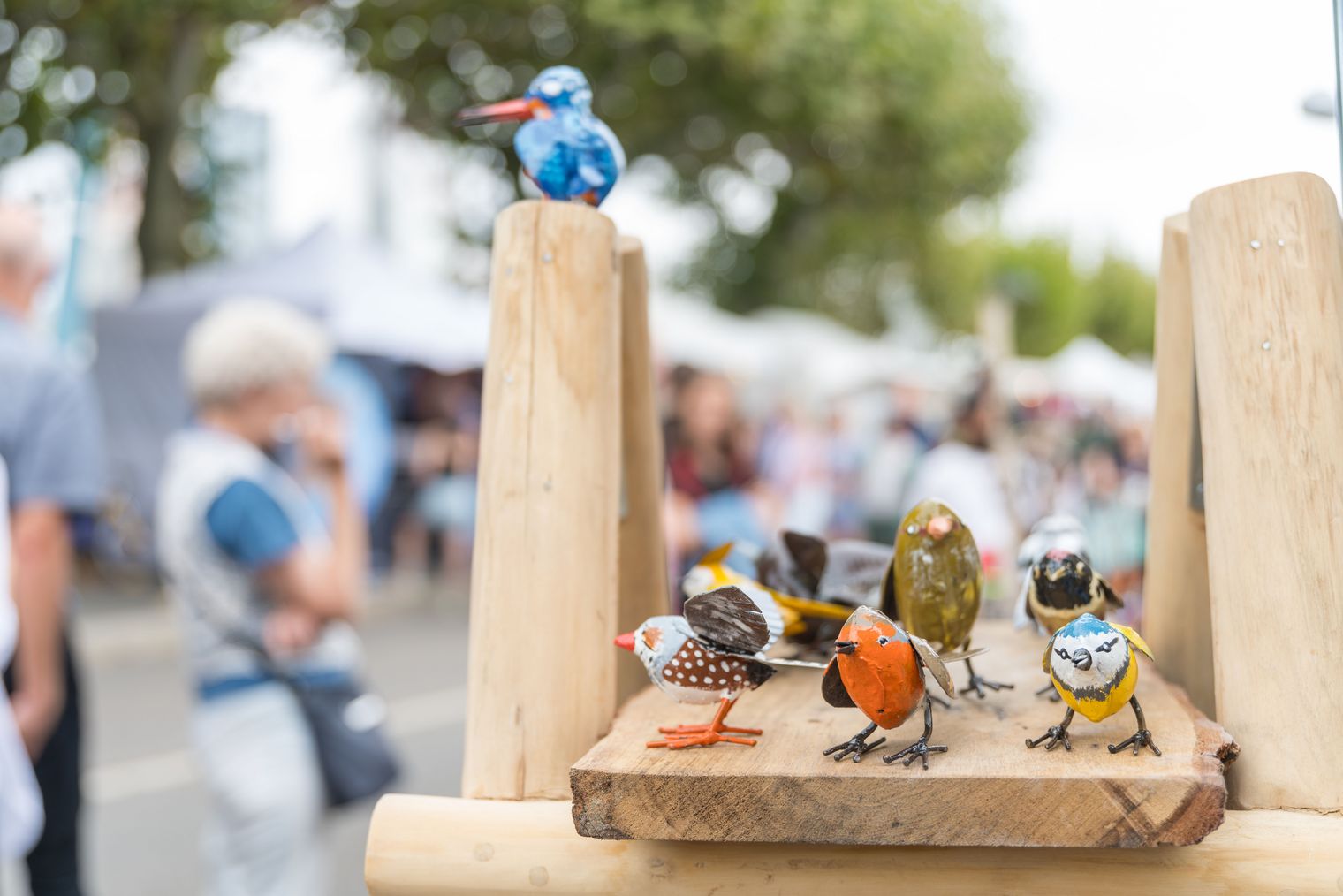 Bemalte Metallvögel an Marktstand mit Besuchern im Hintergrund