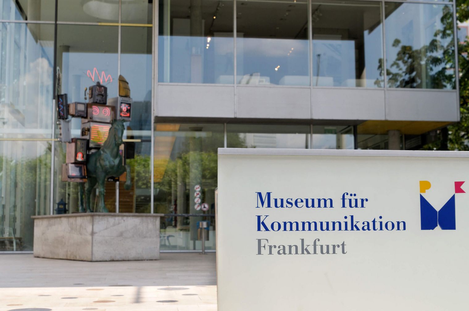 Museumsschild am Eingang des Museums für Kommunikation mit Blick auf den Eingang und eine Metallskulptur