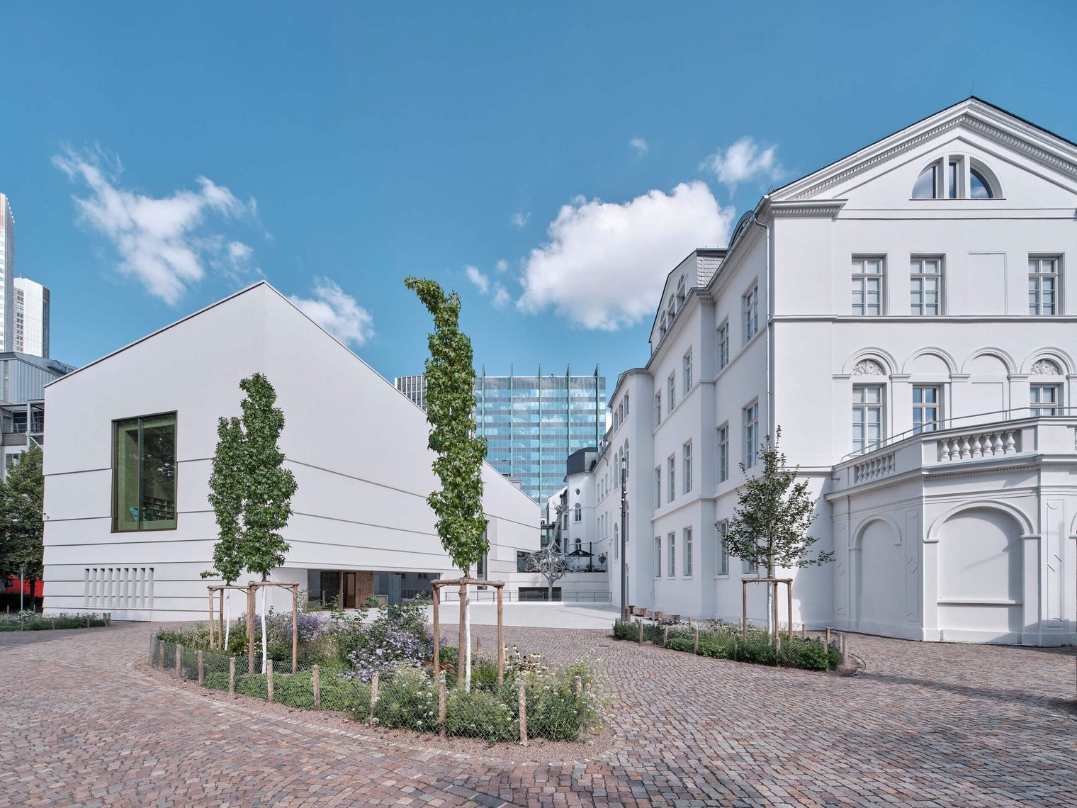 Ein modernes Gebäudeensemble mit weißem Neubau und klassizistischem Altbau, verbunden durch einen gepflasterten Hof mit jungen Bäumen und Blumenbeeten, vor blauem Himmel.