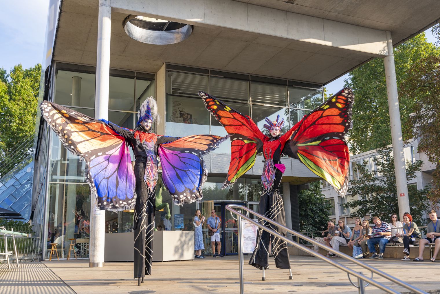 Künstler auf Stelzen mit Schmetterlingsflügeln vor Museum