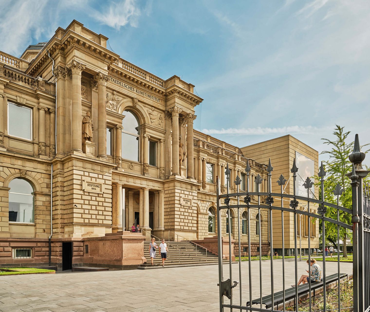 Das Städel Museum in Frankfurt am Main an einem sonnigen Tag. Das historische, neoklassizistische Gebäude mit Säulen und kunstvoller Fassade wird von einem schmiedeeisernen Zaun im Vordergrund eingerahmt. Einige Besucher*innen sind vor dem Eingang zu sehen. Rechts steht ein moderner Erweiterungsbau